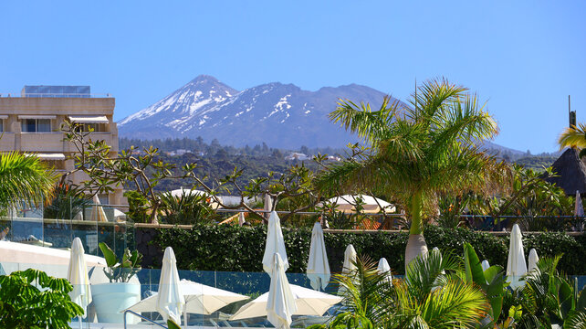 luxury seaside resort on tenerife framed by tall palms and bright umbrellas, opening toward the distant snow‑touched summit of pico de teide rising above the landscape with a calm and expansive island