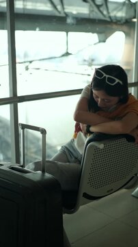 Tired young sola woman traveler sleeping on airport waiting chairs while holding smartphone and boarding pass after long flight delay, bored passenger waiting at airport terminal.
