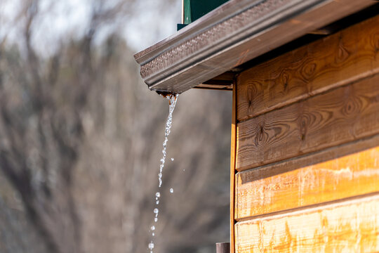 Close-up of water dripping from a brown metal rain gutter on a wooden house in bright sunlight. The image captures the natural process of thawing with a blurred natural background.