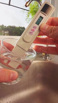 Vertical video. First Person View Of Hand Measuring Drinking Water With Digital Tester Showing Twenty Two Ppm In Glass Near Kitchen Sink And Tropical Landscape Outside Window. Clean Drinking Water Qua