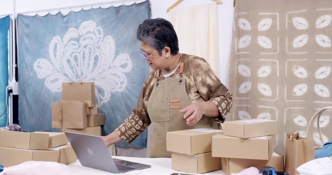 Older asian businesswoman checks order screen in slow motion, sorts parcel boxes by label, confirms shipping address, keeps invoice and receipt ready, home studio e-commerce dispatch continues.
