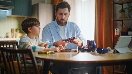 Fototapeta premium Dad help repair toy car little son sitting kitchen watching video tablet closeup