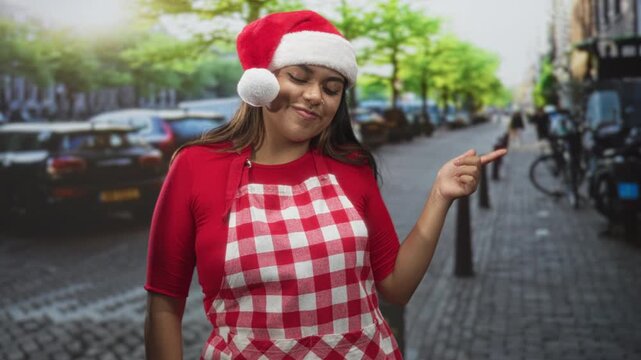 Woman in santa hat and red checked apron points finger on street while smiling with closed eyes; festive joy.