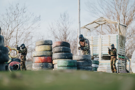 Soldiers in camouflage operate with airsoft guns behind stacked tires. Outdoor training ground shows a tactical setup with cover and wooden structure.