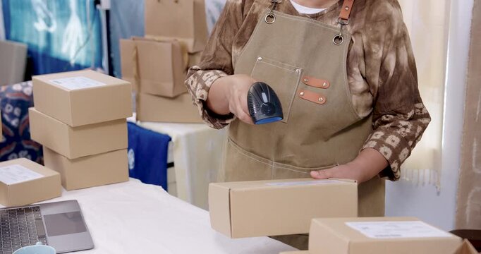  older artisan hands scanning barcode label on parcel box, checks invoice sheet near laptop, rotates carton, verifies order detail, prepares packaging for shipping dispatch home studio.
