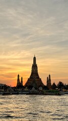 Naklejka premium Wat Arun's grand central prang tower is silhouetted against a vivid orange and golden sunset sky reflected in the Chao Phraya River, with longtail boats moored at the temple's riverside pier. The dram