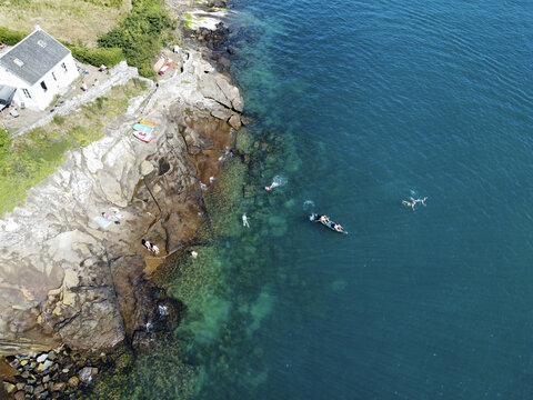 Aerial view of bathers enjoying the clear, turquoise waters beside rugged, rocky shores near a quaint white house, Manche, Normandy, France.