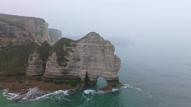 Aerial slow motion tracking pan of chalk cliffs and sea stack eroding with waves on foggy coastline