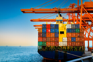 Rear view of a large container ship being discharged by massive orange quay cranes at a port terminal during sunset. © Kosal