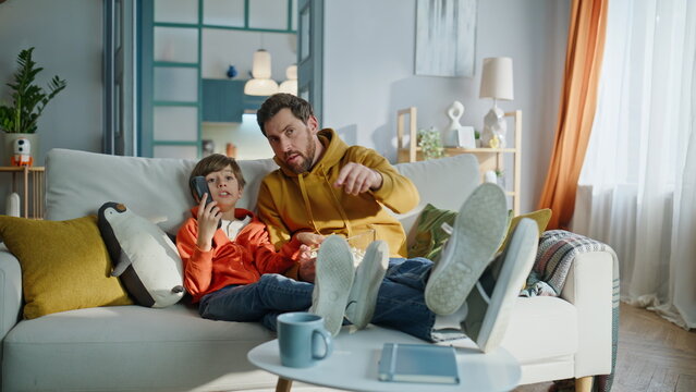 Dad child discussing tv show sitting cozy couch eating popcorn. Cute boy man