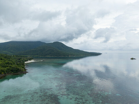 Aerial view of turquoise waters meet lush green hills under a cloudy sky, creating a serene coastal scene at Cottage Legon Waru, Karimunjawa, Central Java, Indonesia.