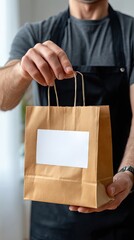 Person holds out empty brown kraft paper bag for food delivery with white label on isolated background wearing black apron and t-shirt