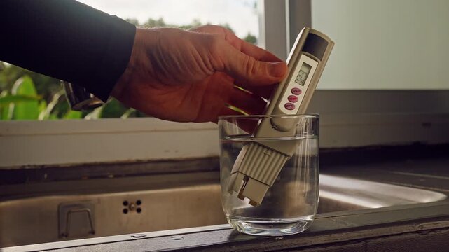 First Person View Of Hand Measuring Purity Of Drinking Water With Digital Tester Showing Twenty Three Ppm In Clear Glass Near Kitchen Sink And Window With Tropical Green Landscape. Water Quality Contr