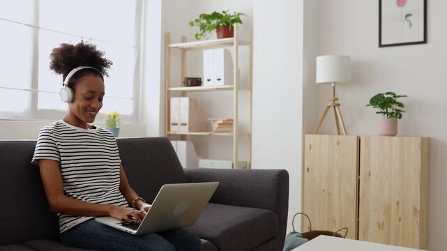 Young smiling african american woman with afro hair listening to music with headphones while typing on her laptop computer, comfortably sitting on the sofa in her cozy living room