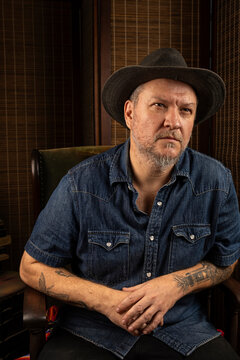 A 48 year old Brazilian man sits in a rocking chair, wearing a hat, with a distant look in his eyes.