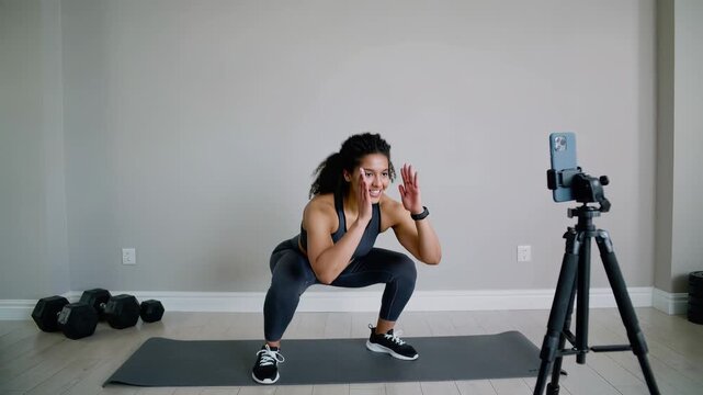 A woman performs a squat exercise in front of a smartphone on a tripod while recording a fitness video at home with dumbbells nearby