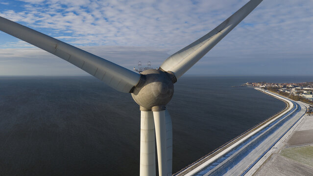 Aerial view of a towering wind turbine, its blades reaching towards the cloudy sky above the calm waters, with a distant coastline, Espel, Flevoland, Netherlands.
