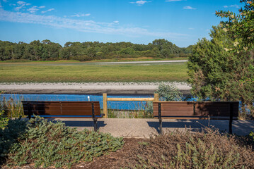 Fototapeta premium Two Park Benches Overlooking Riverbank and Marshland Landscape on a Sunny Day at Bibra Lake Reserve, 1 April 2021, Perth, Australia