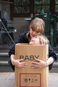 Two sisters share a tender, playful moment as the younger sits inside a cardboard box labeled 'BABY handle with care' while the older embraces her on a porch, conveying warmth, care and childhood fun.