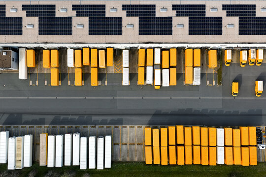 Aerial view of a bustling depot where rows of bright yellow and white delivery trucks stand under a roof of dark solar panels, North Rhine-Westphalia, Germany.