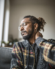 Fototapeta premium A man with glasses and dreadlocks sitting on a couch, holding his neck with a pained expression, wearing a patterned sweater in a softly lit room.