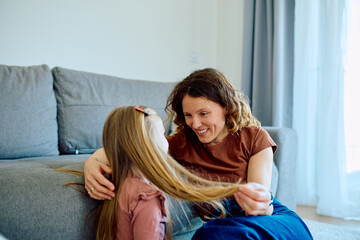 Fototapeta premium Mother and daughter enjoying time together at home, mom smiling and playing with the girl's long blond hair