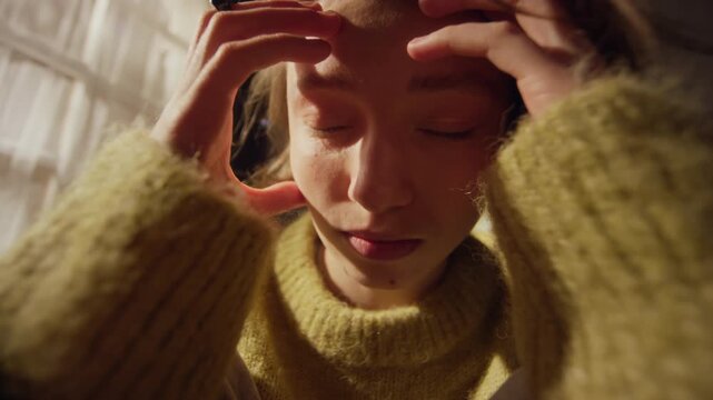 Handheld close up shot of troubled young adult woman sitting in bedroom fidgeting nervously with ring on finger then touching face with hands while experiencing intense anxiety