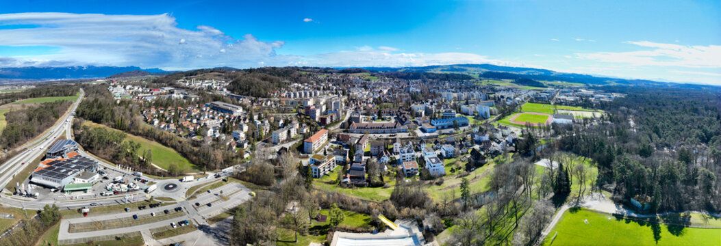 Aerial view of the town nestled amid rolling hills and lush forests under a bright blue sky, Lyss, Canton of Bern, Switzerland.