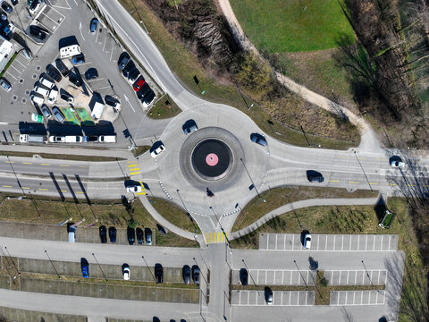 Aerial view of cars navigating the roundabout's concentric circles, framed by parking lots and verdant patches, a dance of motion and stillness, Lyss, Canton of Bern, Switzerland.