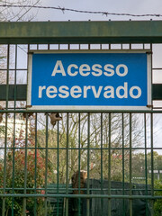 Blue sign reading &ldquo;Acesso reservado&rdquo; mounted on a metal fence with barbed wire, outdoor restricted area warning in Portuguese indicating private access and controlled entry to the enclosed space.