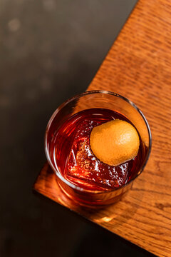 Old fashioned alcoholic cocktail in a transparent rocks glass with a big ice cube and orange peel garnish, lifestyle image on wooden table