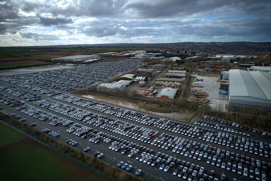Aerial view of row upon row of parked cars contrasting against the industrial buildings and the distant patchwork fields under a brooding sky, Stone, England, United Kingdom.