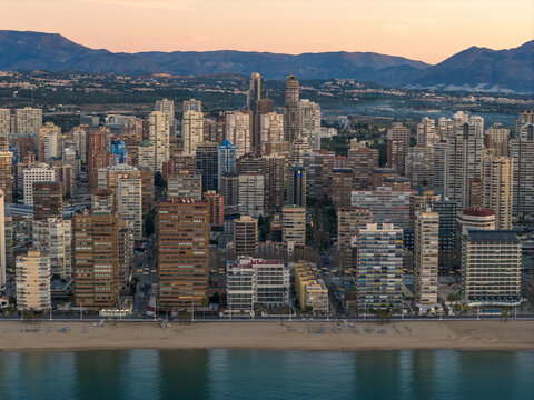 Aerial view of the skyline where golden sands meet the turquoise sea, overshadowed by towering buildings against a backdrop of distant mountains, Benidorm, Valencian Community, Spain.
