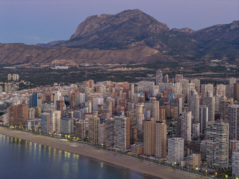 Aerial view of towering skyscrapers meet the tranquil sea, shadowed by majestic mountains, as the city lights begin to twinkle, Benidorm, Valencian Community, Spain.