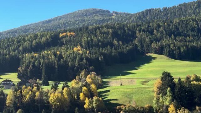Scenic drive through Rasun-Anterselva valley in the Dolomites, Italy. View from car window of green alpine meadows, forested hills and sunlit mountains under clear blue sky. Horizontal video.