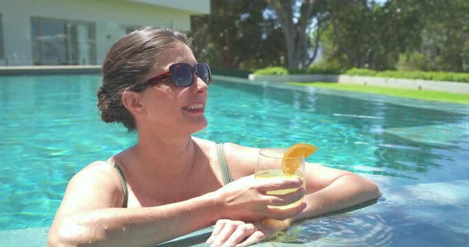 Mature adult female relaxing in sunlight at tiled pool edge, sipping drink with orange slice