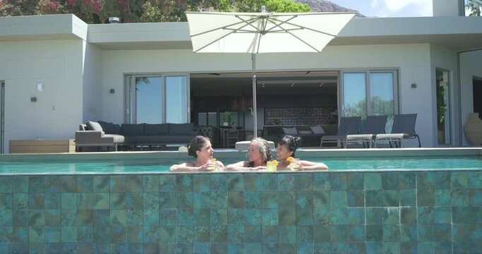 Diverse female friends in swimsuits relaxing chatting with drinks as camera closing in at pool