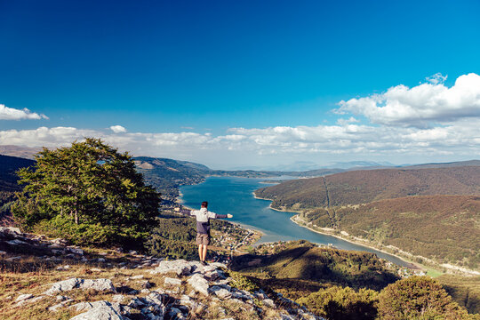 View of a man standing on a rocky outcrop, arms outstretched, overlooking a serene lake nestled between rolling hills under a vibrant blue sky, Mavrove, North Macedonia.