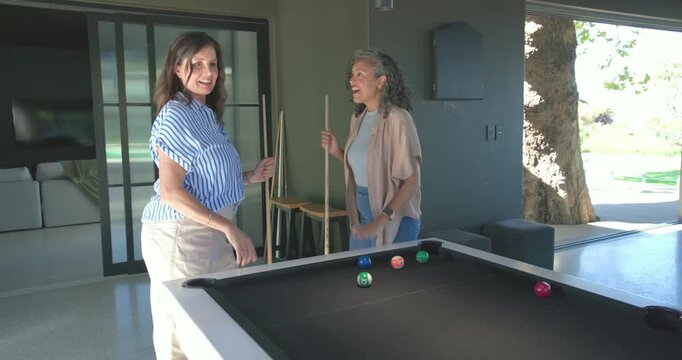Diverse female friends holding cues, facing pool table at home, pointing to balls, planning shot