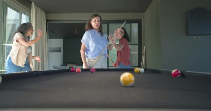 Diverse female friends playing pool in rec, watching right woman striking yellow ball on black felt