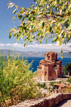View of an old church standing proudly on a hill, overlooking the serene blue waters under a clear sky, framed by delicate tree branches, Ohrid, North Macedonia.