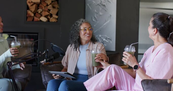 Diverse women cohosts on record cue passing green smoothies adjusting mic moderating tablet podcast