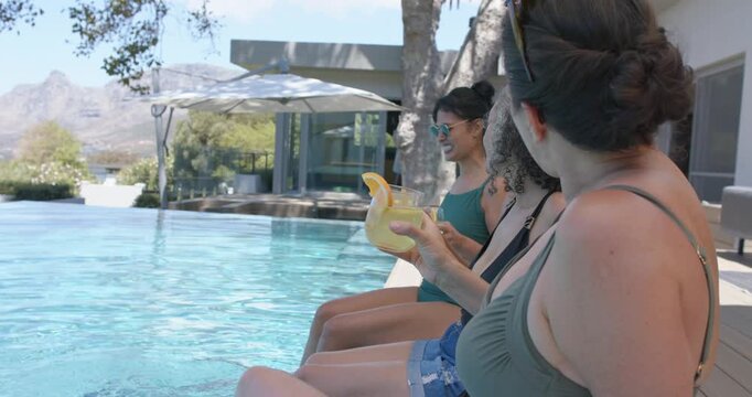 Diverse female friends chatting at home pool edge, middle woman speaking with yellow drink