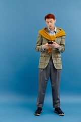 Naklejka premium Stylish young man checks smartphone while dressed in trendy attire against blue backdrop