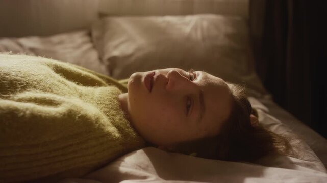 Portrait shot of young adult woman lying down on bed in sunlight then turning to camera with distressed expression while struggling with emotional crisis