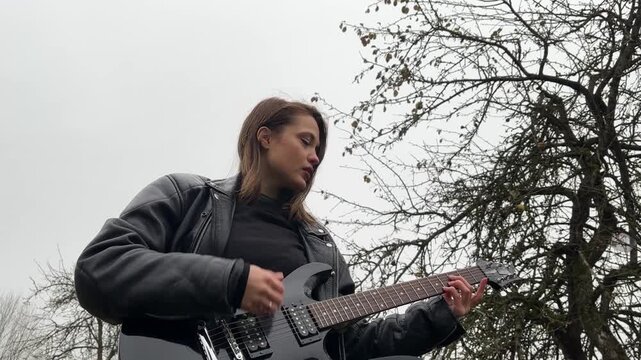 A female rock musician in a leather jacket plays an electric guitar with an amplifier in the garden in gloomy weather