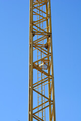 A tall yellow industrial tower crane mast with internal ladders stands vertically against a solid clear blue sky in bright daylight