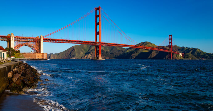 Wide-angle panorama of the world-famous red Golden Gate Bridge on a windy, sunny spring morning, viewed from Torpedo Wharf on the south side. Popular tourist attraction and landmark in California, USA