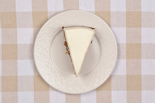 A top down view of a triangular slice of chocolate cream cake with white frosting served on a decorative plate over a beige and white checkered fabric background