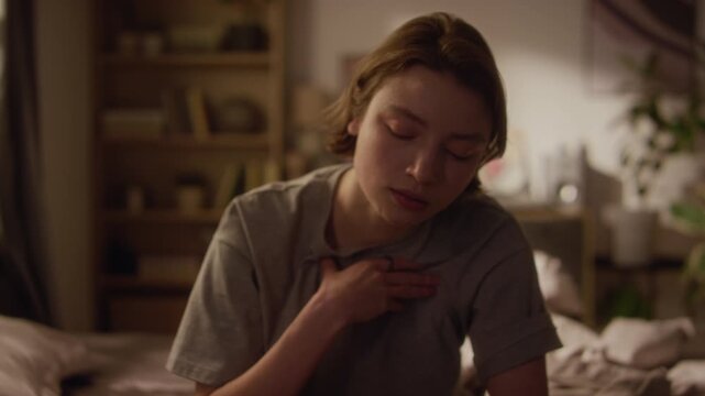 Tilt up shot of young adult woman touching chest feeling heart pounding while having panic attack in dimly lit bedroom